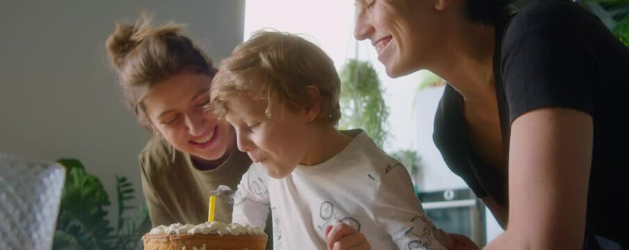 Joyful lesbian mothers kissing little son as he blowing candle on birthday cake during family celebration at home