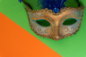 image of a blue and gold venetian mask with blue feathers on and black ribbons orange and green background with copyspace