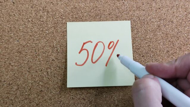 Handwritten inscription "50%" on a yellow paper sticker. Writing with a red felt-tip pen. A woman's hand with a colored marker close-up. Sticker on a cork board