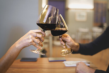 Couple toasting red wine glasses sitting at restaurant table - close up on the hands and clinking wineglasses - dating concept