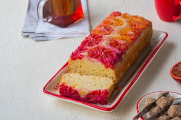 A whole cake with blood oranges on top on a white rectangular plate on a light concrete background.