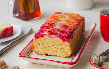A whole cake with blood oranges on top on a white rectangular plate on a light concrete background.