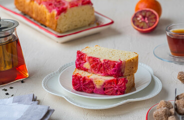 A sliced cake with blood oranges on top on a white plate on a light concrete background.