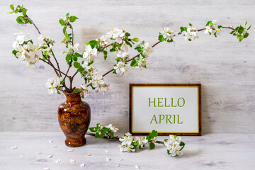 Spring still life with branches of blossoming apple tree in vase with photoframe on light background. White hotoframe with text HELLO APRIL. April is coming, springtime concept