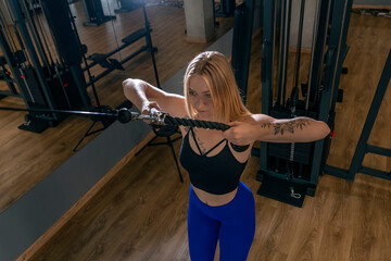 Fototapeta na wymiar Fitness woman trains triceps muscles in cable crossover exercise machine. Blonde girl with beautiful figure in the gym.