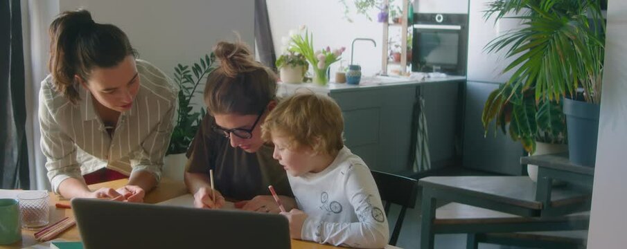 Young lesbian parents helping their little son with doing homework while sitting together at table in kitchen at home