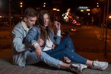 Young caucasian couple is sitting on the pavement embracing against the backdrop of the lights of the night city
