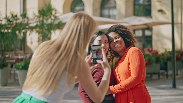 Blond woman taking photos of happy lesbian couple posing outdoors, love