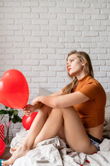 Beautiful thoughtful woman holding red heart shape balloon sitting in bed