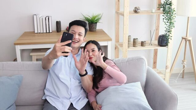 Young Asian couple say hi and talking in video conference by smartphone together in living room at home, Activity with technology lifestyle on mobile phone concept