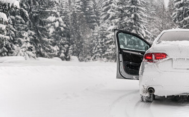 Silver car parked on snow covered winter road, front door open, blurred trees background, view from behind