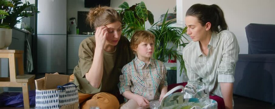 Lesbian parents and little son sitting on floor at home, chatting and sorting waste into different bags while preparing it for recycling