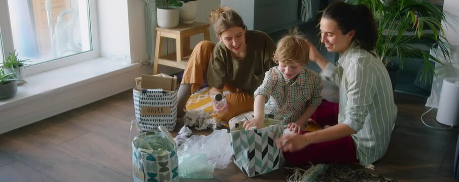 High angle shot of cheerful lesbian mothers and little son sitting on floor at home and sorting waste into different bags while preparing it for recycling