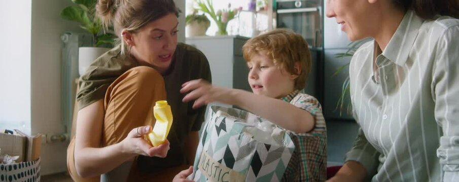 Cute little boy sitting on floor at home and collecting plastic into bag while helping lesbian parents with sorting waste for recycling