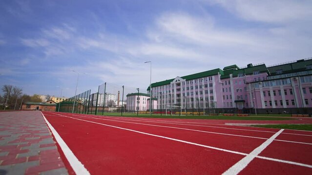 Approaching a soccer field surrounded by fence in front of the big pink building. Red running track at the foreground. Low angle view.