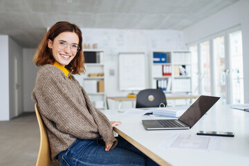 young business woman with glasses sits at a desk with laptop and laughs into the camera