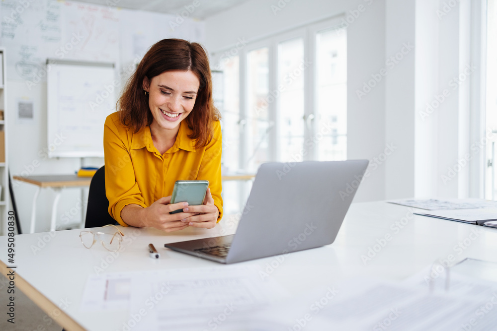 Wall mural young modern business woman sitting at desk and looking at her cell phone in office