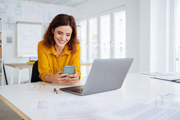 young modern business woman sitting at desk and looking at her cell phone in office