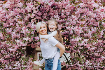 People, holidays and family concept - daughter hugs mom sitting on her back on cherry blossom background. Walk in the sakura trees in the city.