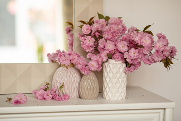 Three vases with sakura twigs in beige vases in the home interior. Mirror at the window in the dressing room.