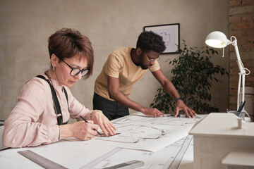 Concentrated mature woman in glasses sitting at table and creating drawing for architectural project together with African-American intern