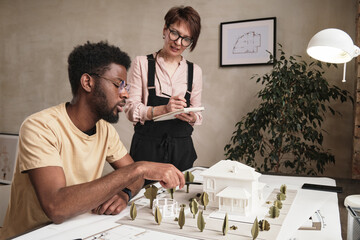 Young African-American architect in glasses pointing at D model and discussing landscape design with colleague