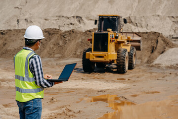 A young man civil engineer working at sand quarry inspects the operation of yellow excavators and...