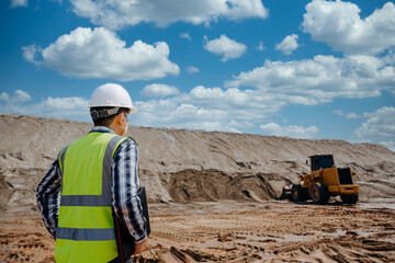A young man civil engineer working at sand quarry inspects the operation of yellow excavators and...