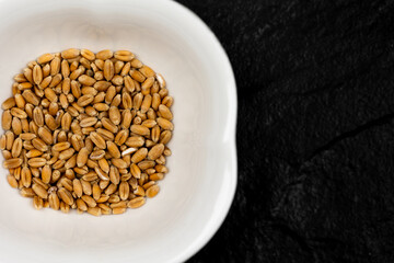Top view of wheat grain in white bowl standing on black, stone surface. Scene lit with artificial, soft light.