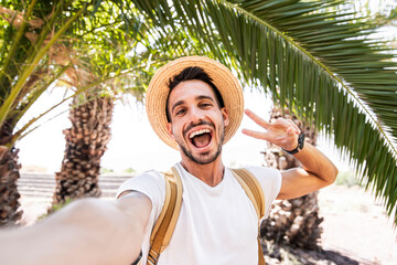 Young man with backpack taking selfie portrait on exotic beach -Smiling happy guy enjoying summer holidays - Millennial showing victory hands symbol to the camera - obrazy, fototapety, plakaty