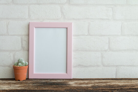 Blank pink photo frame on old wooden table with cactus over white brick wallpaper background copy space. Lifestyle and home decoration concept.