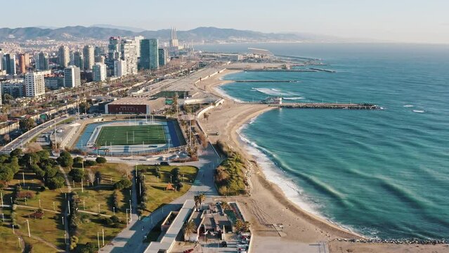 Extreme wide aerial shot of densely populated big modern city located on sea cost. Camera flying above residential areas, roadways, park and stadium to sandy seashore bathed in waves. Barcelona, Spain