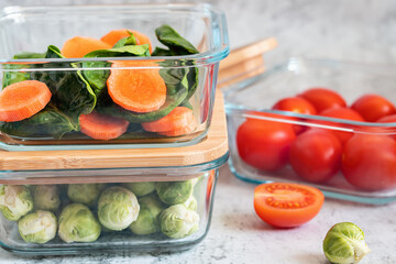 Various vegetables in glass containers: carrots, spinach, tomatoes, Brussels sprouts. Vegan food and snacks in containers, gray background. Clean eating, raw food, detox,
