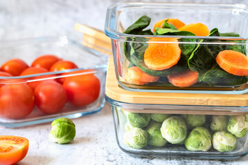 Various vegetables in glass containers: carrots, spinach, tomatoes, Brussels sprouts. Vegan food and snacks in containers, gray background. Clean eating, raw food, detox,