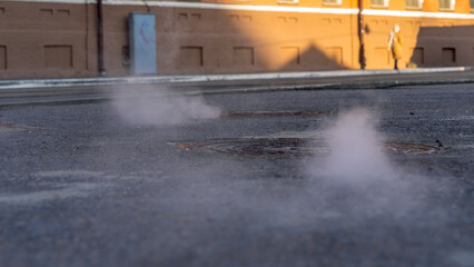 Manhole cover in a paved street on a cold day, as steam escapes from the sewer below up into the city street.