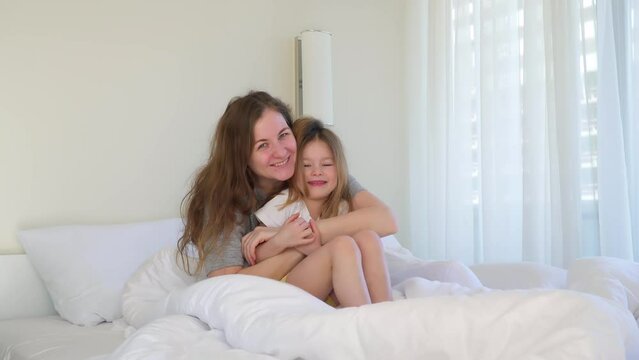 Cuddling in bed. daughter hugging a woman for mother's day. cute little girl sits under blanket with white pillows. hotel room is bright and white. Caucasian sisters have fun together. family smiles.