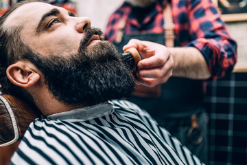 The middle-aged barber trims a young guy's beard in his barber shop.
