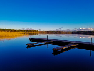 a wooden pontoon for boats stands on the water of the lake and is reflected in the water. In the distance, the forest and reeds are reflected in the water of the lake.