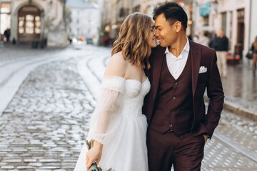 Naklejka premium An international wedding couple, a European bride and an Asian groom walk around the city together.