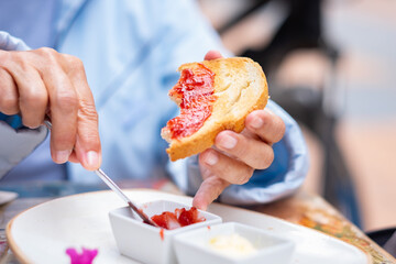 Mature woman holding slice of toast putting strawberry jam on top while sitting at an outdoor cafe...