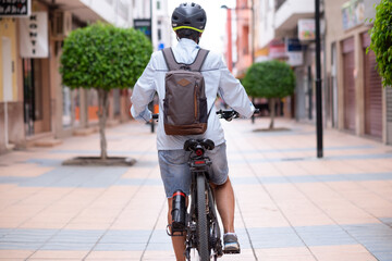Naklejka premium Rear view of senior cyclist woman wearing helmet on electric bicycle in city street looking ahead. Active elderly grandmother enjoying a healthy lifestyle and freedom