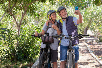 Obraz premium Active caucasian senior couple with electrobikes standing outdoors in the park video calling by cellphone. Two smiling elderly people wearing helmet running in nature enjoying healthy lifestyle