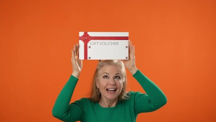 Happy mature blonde woman 40s 50s dancing with gift certificate voucher thumbs up winner gesture isolated on orange color background studio portrait.