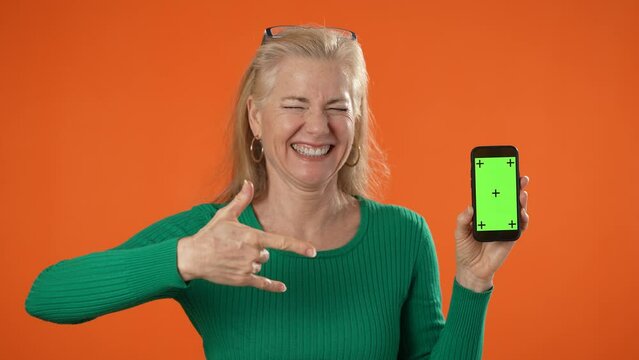 Smiling mature woman posing holding phone with tracking points on blank empty green screen, isolated on orange background. Giving ILY I Love You hand sign. 
