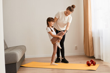 Fototapeta na wymiar Young woman with little girl wearing sportswear doing sport exercises at home, kid standing with one leg lift, thighs workout, warming up training muscles, child having workout with trainer.