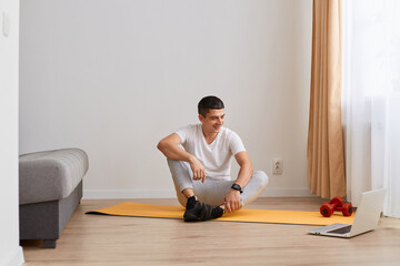 Horizontal shot of smiling Caucasian man wearing sportswear doing sport exercises at home, sitting on floor on yoga mat, looking at laptop screen, listening instruction online before workout.