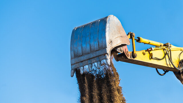 Loose soil falling down from excavator shovel lit by spring sunlight on a blue sky background. Close-up of yellow digger arm metal bucket when dumping brown dirt. Working hydraulic earth mover detail.