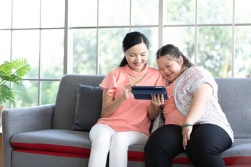 Obraz premium mother using tablet computer with a girl down syndrome or her daughter, smiling and enjoying on sofa