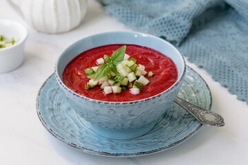 Beet and Tomato Gazpacho. Bowl of ruby red cold gaspacho puree soup topped with cucumber cubes and fresh basil on light marble background.