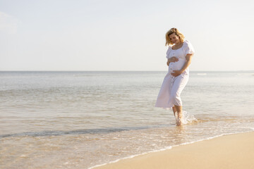 Beautiful pregnant woman in white dress walking barefoot on a beach
